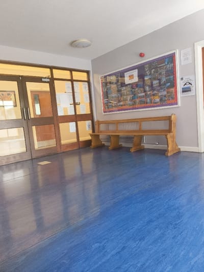 The image shows the interior of a building located in Kilkeel, United Kingdom (latitude: 54.065, longitude: -6.031). The building is brightly lit with a single light fixture on the ceiling. The room features a blue tiled floor, a wooden bench with a back and arms, and a set of double doors leading to an adjacent room. The double doors are wooden and have glass panels. The glass panels are covered with paper, presumably containing handwritten notes or schedules. A large bulletin board is positioned on the wall, displaying a collage of images. The bulletin board is mounted above the wooden bench. The walls are a pale grey color. There are no people present in the room. The room appears to be empty and waiting for occupants.