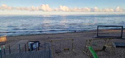 This image depicts a coastal scene in Vārzas, Latvia, under a clear sky with scattered clouds. The foreground features a sandy beach with several recreational elements. To the left, a wooden deck with railings overlooks the beach and water. Further to the right on the sand, there are two swings, one with a circular rope seat and another with a wooden plank seat, both made of bamboo frames. Adjacent to the swings is a small green slide attached to a wooden structure. In the background, a large, dark rectangular frame stands on the beach, with the word "VĀRZAS" visible on its upper beam. Beyond the beach, a calm body of water stretches towards the horizon, with gentle waves lapping the shore. The water reflects the blue of the sky and the white of the clouds. No people are visible in the image.