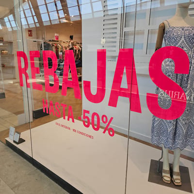 This is a photograph taken through a glass storefront, likely a clothing store, in Zaragoza, Spain. The most prominent subjects are large, bright pink lettering applied to the glass, advertising a sale. The lettering is partially obscured by reflections of the interior of the store and the exterior environment.

The words visible are "SALE" in large letters, and "HASTA 50%" which translates to "UP TO 50%". Below this, the text "EN EL INTERIOR VER CONDICIONES" is visible, meaning "INSIDE SEE CONDITIONS".

Inside the store, visible through the glass, are mannequins dressed in clothing. One mannequin, partially visible at the top of the frame, wears a blue and white patterned dress and beige sandals. Further into the store, a person, likely a staff member, is arranging clothing on racks. There are also signs indicating discounts, such as "-20%" and "49". The interior appears to be a well-lit retail space with wooden flooring and modern fixtures.

The reflections on the glass show a grid of windows, suggesting the store is located within a mall or a building with a large glass facade. The lighting suggests it is daytime. The overall scene conveys a commercial environment focused on attracting customers with a sale.