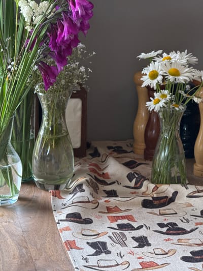 This is a close-up, eye-level, indoor shot of a rustic table setting. In the foreground, a light-colored fabric runner is spread across a wooden surface. The runner is adorned with a repeating pattern of cowboy hats, boots, and cacti in shades of brown, black, and orange.

To the left, a clear glass vase holds a bouquet of tall, thin green foliage and delicate white flowers. Beside it, another clear glass vase, wider at the base and narrowing towards the neck, contains a spray of vibrant purple gladiolus flowers and more small white filler flowers.

To the right of the purple flowers, a vase with a similar shape to the one holding the gladiolus contains a bouquet of white daisies with yellow centers. Behind the daisy bouquet, three wooden salt and pepper grinders are visible. The grinder on the left is a lighter wood, the middle one is dark brown, and the one on the right is a darker shade of brown or black. A dark, rectangular object, possibly a napkin holder with a decorative wood frame, stands behind the purple flowers.

The background is a plain, dark gray wall. The overall scene evokes a western or country aesthetic. There are no people visible in the image. The lighting suggests an indoor setting, possibly with natural light coming from the left, casting subtle shadows.

While the image depicts a table setting, its specific location within the city of Dsegh, Armenia, cannot be determined from the visual information provided.