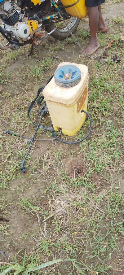 In Angwan Hausawa, Nigeria, a yellow plastic sprayer with a blue lid rests on muddy ground covered in dry grass and sparse green vegetation. A black hose connects to the sprayer and extends to a spray wand with a red nozzle tip. Behind the sprayer, a motorcycle is partially visible, featuring a white engine block and a yellow tank mounted over the rear wheel. The motorcycle has colorful blue, yellow, and green stripes on its frame. Bare feet are visible on the ground near the motorcycle, one of which is dirty. The overall scene depicts an outdoor setting, likely agricultural or rural, with evidence of recent use or storage of farming equipment.