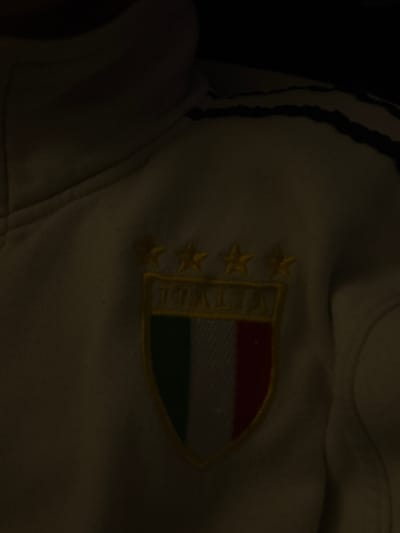 The image shows a close-up of a white garment with an embroidered Italian national team badge. The badge is shield-shaped and features the colors of the Italian flag (red, white, and green) in horizontal stripes. The word "ITALIA" is embroidered vertically along the left side of the shield. Three gold stars are located to the left of the text. The lighting is poor and doesn't allow for a detailed description of garment's details. The location of Menenkyovo, Bulgaria is not discernible from the image.