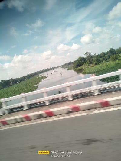 The image shows a blurred, diagonal view from a moving vehicle, likely a car, looking out over a river and its banks. In the foreground, a white railing with red and white stripes on its base is visible, suggesting the perspective is from a bridge or elevated walkway. The river itself is wide and calm, with sections covered by dense green aquatic plants, possibly water hyacinths, forming a thick carpet on the water's surface. The far bank of the river is lined with lush green trees and vegetation, with some buildings and structures faintly visible in the distance. The sky is partly cloudy, with a mix of white and wispy clouds against a pale blue background. The timestamp "2026.06.24 09:59" and the watermark "realme Shot by zsm_travel" are present in the lower right corner, indicating the image was taken with a Realme phone and its date and time. The overall impression is a scenic view of a natural landscape along a waterway, likely captured during daylight. The location is identified as Gò Dầu, Vietnam.
