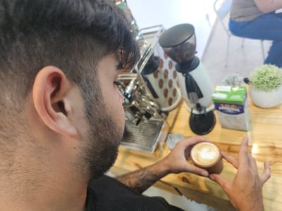 A man is seen in profile, with a focus on his ear and facial hair. In his hands, he holds a small cup of coffee with latte art. Behind him on a wooden counter is a coffee grinder, a carton of milk, and a potted green plant. In the background, a metal coffee machine and a person seated on a chair can be seen. The scene suggests the man is a barista or enjoying coffee preparation.