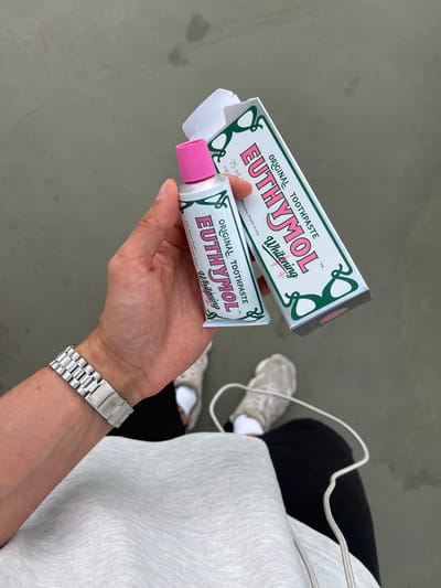 This media file features a close-up view of a person's hands holding a box and a tube of Euthymol Original Toothpaste. The toothpaste box is primarily white with green decorative accents and bold pink lettering that reads "EUTHYMOL." The tube, also white with similar branding, has a pink cap. The text "ORIGINAL TOOTHPASTE," "Whitening," and "Whitening Double Action" is visible on both the box and the tube. The person is wearing a silver wristwatch with a metal band. In the background, the lower body of another person is visible, wearing grey athletic shoes, black pants, and a light grey sweatshirt or hoodie. The scene appears to be outdoors, with a grey, textured ground surface. The lighting suggests it could be daytime. There are no strong indicators of weather conditions. The context provided indicates the location as Borowa, Poland. The overall impression is one of a casual, everyday moment, possibly shopping or preparing for a trip, with a focus on the iconic Euthymol toothpaste.