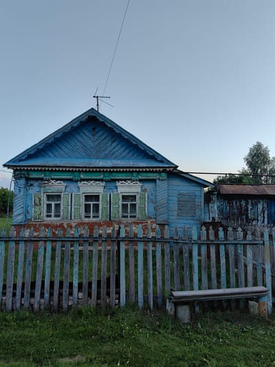 A worn, two-story wooden house painted blue with peeling paint stands behind a weathered, blue-painted wooden fence. The house has three windows on the upper floor, each with green shutters. A decorative wooden trim runs along the top of the house. A small, dilapidated shed with a rusted roof is attached to the side of the house. In front of the fence, a concrete bench sits on the grass. The sky is clear and pale blue, indicating it is either early morning or late evening. The location is in Krestovo-Gorodishche, Russia.
