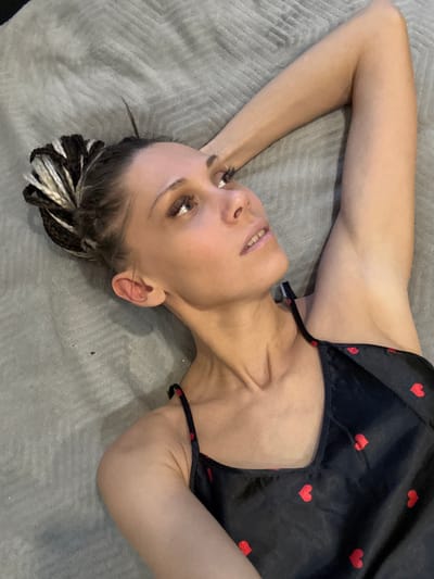 A woman is lying on a gray surface. She is wearing a black top with red hearts. Her arms are above her head, and she is looking up. Her hair is styled in black and white braids. The location is unspecified, and there is no evidence to suggest the location is Yaroslavl', Russia.