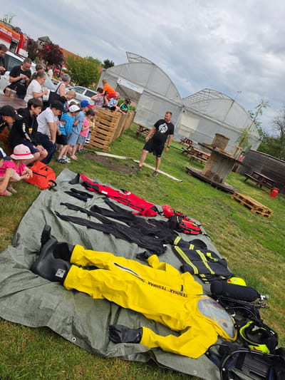 The image shows a scene in Mlčechvosty, Czechia. A group of people, mostly children, are seated on a bench, facing a presentation of firefighting equipment. The equipment is laid out on a green tarp on the grass. It includes a yellow hazmat suit, black and red fire-resistant clothing, helmets, and oxygen tanks. A man in a black t-shirt and shorts is standing near the equipment, possibly giving the presentation. In the background, there are greenhouses and wooden structures. A red fire truck is visible on the left side of the frame. The sky is cloudy.