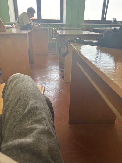 The image depicts an indoor scene, likely a classroom or study hall, with wooden desks and chairs. In the foreground, a person's legs are visible, clad in grey trousers. The person appears to be sitting at a desk. Further back, another individual, a young male with light hair, is seated at a desk, looking at a mobile phone. He is wearing a light-colored t-shirt and a dark bag is slung across his chest. The room has large windows that allow natural light to enter, illuminating the space. The floor is a reddish-brown tiled surface. The overall impression is that of a quiet, everyday moment within an educational or study environment. The location is identified as Жыровіцкі сельскі Савет, Belarus.