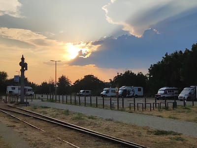 The image shows a sunset scene with several RVs parked in a lot next to a set of train tracks. The sky is filled with dramatic clouds, with the sun peeking through and casting rays of light. The RVs are varied in size and design, and some appear to be unoccupied. The train tracks are in the foreground, with dry grass and vegetation on either side. In the background, there is a line of trees. A sign that reads "CAMINO NATURAL" is visible on a pole near one of the RVs. The overall impression is one of a tranquil, somewhat rustic setting during the golden hour. This scene is likely located near the train station in Béjar, Spain, given the presence of railway infrastructure and the "Camino Natural" sign, which often indicates routes through natural landscapes in Spain.