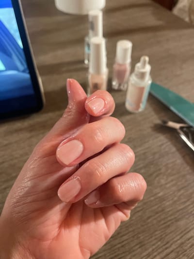 A close-up, slightly elevated shot captures a person's left hand resting on a wooden surface in Monsummano Terme, Italy. The hand displays five fingernails, each coated with a translucent, light pink or nude-colored nail polish. The skin on the fingers and palm exhibits a distinct sheen, indicative of recent application of moisturizer or cuticle oil. In the blurred background, positioned on the same wooden surface, are various nail care items: four small bottles with white caps, likely containing nail polishes or treatments, including one with a dropper applicator. To the right, a teal-colored nail file and a metal cuticle nipper are present. A dark electronic tablet or smartphone, with a blue-toned screen, is partially visible on the far left of the frame.
