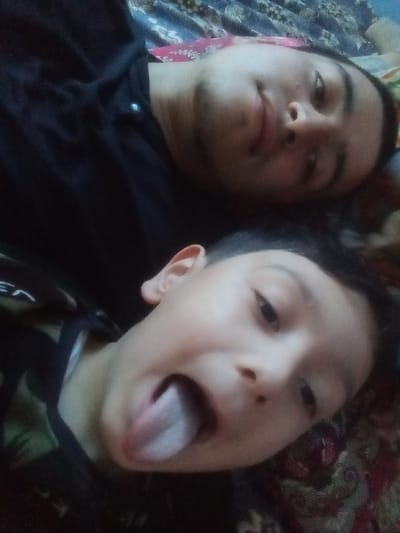 The image depicts two males lying down in what appears to be an indoor setting, possibly in Kasan, Uzbekistan. An adult male is visible in the upper portion of the frame, positioned on his back. He has short dark hair and visible facial stubble, wearing a dark collared shirt with a single button near the collar. His expression is neutral with a slight upward turn at the mouth. Below him, a younger male, identified as a child, is also lying down, facing the camera. The child has short dark hair and is wearing a dark shirt with a subtle pattern on the shoulder. His mouth is open, and his tongue is extended outwards, indicating a playful action. Both individuals are looking directly towards the camera. They are situated on a patterned fabric surface, consistent with a blanket or bedding, which displays floral or abstract designs in shades of blue, red, and brown.