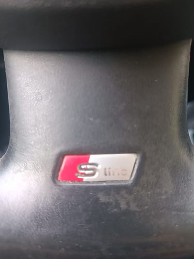 This media file is a close-up, slightly angled shot of the center of a car's steering wheel, specifically focusing on the "S line" emblem. The emblem is metallic with a silver "S" and the word "line" also in silver, set against a background split between a bright red and a brushed silver rectangular area. The steering wheel itself is dark and appears to be made of leather or a similar material, showing some texture and possibly light wear. The background is dark and out of focus, suggesting the interior of a car. The lighting is moderate, with some glare on the metallic emblem. There are no people visible, and the activity is simply the stationary presence of the steering wheel. The location context provided is Germersheim, Germany, which is not visually indicated in the image itself but serves as contextual information. The text "S line" is clearly visible on the emblem.