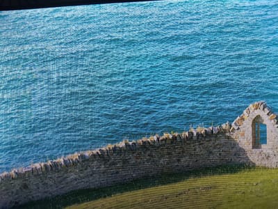 The image shows a stone wall with a ruined structure on the right side. The ruined structure has an arched window with panes. The wall appears to be bordering a grassy area. The background is a body of water with visible waves. The scene is viewed from an elevated perspective. The image is a photograph, and appears to be digitally captured as evident by the pixelation. There are no people or animals visible in the image. The location is not in the city of Reftinsky, Russia.