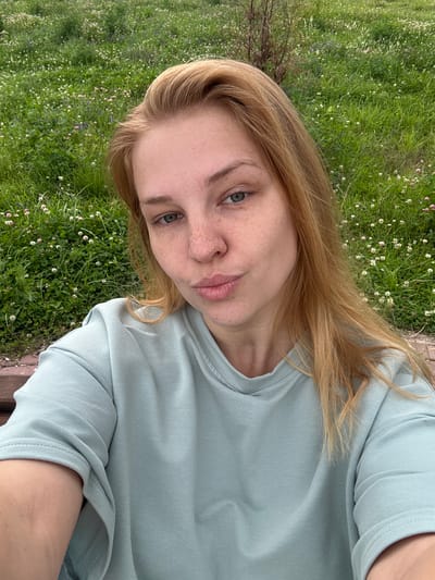 The image is a selfie of a young woman with fair skin, blonde hair, and blue-green eyes. She has freckles across her nose and cheeks. Her lips are slightly parted, and she is looking directly at the camera with a neutral expression. She is wearing a light blue, long-sleeved t-shirt.

The background is a grassy area with small white and pink wildflowers, suggesting a meadow or park. The lighting indicates it is daytime, likely with overcast or soft lighting given the absence of harsh shadows. The scene has a natural, outdoor feel, possibly in a rural or park setting.

The orientation of the image is rotated, with the woman appearing to be lying down or leaning back. The overall impression is of a relaxed, casual moment captured outdoors. There is no visible text in the image.