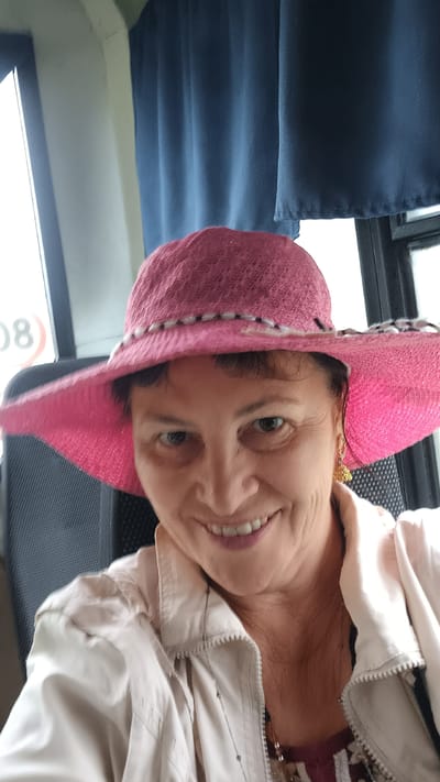 A medium shot, taken from a slightly low angle, shows a woman wearing a pink sun hat and a light-colored jacket. The woman has medium-length brown hair and is smiling at the camera. Her earrings are visible, and she is wearing a necklace. The background consists of a window with blue curtains and a glimpse of what appears to be the exterior of a vehicle or building. The woman is seated, with a portion of a dark-colored chair visible to her left. The overall lighting suggests an indoor setting, possibly during daylight hours. The location is not specified in the image itself.