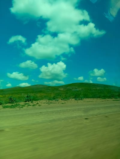 The image depicts a landscape scene with rolling green hills under a bright blue sky dotted with white, fluffy clouds. In the foreground, there is a dirt road or path with some dry grass and small bushes. The overall impression is of a rural or natural environment, possibly viewed from a moving vehicle due to the motion blur in the foreground. The location is specified as Aleksandrovka, Kazakhstan. There are no people or specific actions visible in the image. The scene is characterized by its natural elements: sky, clouds, hills, and vegetation.