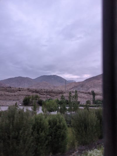 The image shows a view of the mountainous landscape and vegetation in Upshi, India. In the foreground, there is a dark vertical object on the right side, possibly a pillar or frame, obscuring part of the scene. Beyond this, a body of water, likely a river or stream, is visible. Dense green trees and bushes line the banks of the water. In the middle ground, the terrain transitions to a rocky, barren landscape with layers of sediment. Further in the background, rugged mountains rise against a cloudy, overcast sky. There are no people or any discernible actions or interactions visible in the image. The overall impression is of a natural, somewhat desolate environment.
