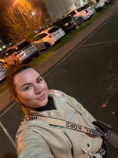 A woman stands on what appears to be the edge of a sidewalk or walkway in an urban environment. She is wearing a light-colored leather jacket and a bag with a shoulder strap labeled "MICHAEL KORS". Behind her is a row of parked cars along the curb, and the scene is dimly lit, suggesting it is nighttime. The location appears to be the city of Proletarskii, Russia.