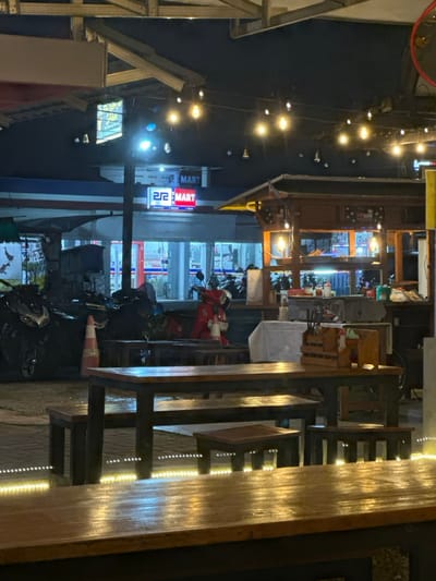 The image depicts an urban street scene at night, likely in Kecamatan Cisaat, Indonesia, illuminated by various artificial lights. In the right foreground, there is an outdoor seating area featuring robust wooden tables and chairs. The tables are distinctly lit from underneath by warm LED strip lights, creating a bright, striped pattern on their surfaces.
A string of warm, glowing bulb lights hangs overhead, stretching across the frame against the dark night sky, contributing to a cozy ambiance. In the mid-ground, a building with a traditional-looking dark roof and an illuminated interior appears to be a commercial establishment. Parked in front of this building are several motorcycles and scooters, including a prominent red one, along with a visible orange traffic cone.
Further in the background, a brightly lit storefront is visible. A prominent sign, predominantly red and blue, clearly displays the text "MART" and "22 MART," with the "22" in a distinct blocky font. The windows of this establishment show reflections and the interior, likely stocked with various goods.
The scene is devoid of people, suggesting a quiet moment, possibly late in the evening or early morning. The combined effect of the string lights, storefront illumination, and table lighting creates a warm and inviting atmosphere. The architecture, motorbikes, and "MART" sign are consistent with a commercial area in Southeast Asia.