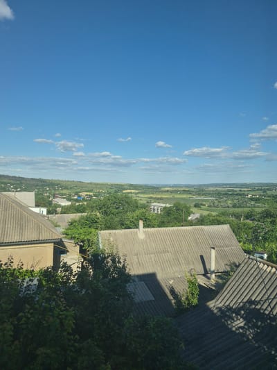 The image captures a panoramic view of a rural settlement in Merenii Noi, Moldova, under a clear blue sky dotted with scattered white clouds. In the foreground, several houses with corrugated asbestos cement roofs are visible, some partially obscured by lush green trees and bushes. One house in the foreground has a tan-colored facade and a visible yellow pipe extending along its exterior wall. Chimneys are present on some of the roofs.
Beyond the immediate houses, the landscape unfolds into rolling green hills and fields, characteristic of the Moldovan countryside. Smaller buildings and houses are scattered throughout the midground, interspersed with dense patches of trees and vegetation. In the distance, the terrain becomes softer and more undulating, with a hazy horizon. The overall scene depicts a peaceful, somewhat rustic residential area integrated with the natural landscape. There are no people or vehicles visible in the image.