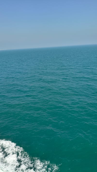 The aerial image displays a transition from a light blue, hazy sky to a deep teal ocean. The sky occupies the left third of the frame, appearing calm and vast. As the image moves to the right, the sky gradually merges into the ocean, creating a soft, blended horizon. The ocean itself is characterized by gentle ripples and waves that create a textured surface. The water's color deepens from a lighter blue-green near the sky to a rich, vibrant teal on the right side.
In the upper right quadrant of the ocean, there are white frothy waves, suggesting movement, possibly from a boat or the sea itself. There are no visible people or identifiable objects in the main body of the water. The scene is serene and depicts a natural seascape. The lighting suggests it could be daytime, with the sun illuminating the water and creating subtle highlights on the waves. There are no textual elements visible in the image.