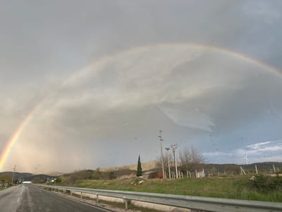 The image depicts a road scene in Bogdanci, North Macedonia, under an overcast sky. A full rainbow arches across the sky. The road is wet, and a metal guardrail runs along the right side. Beyond the guardrail is green vegetation and a landscape with trees, shrubs, power poles, a small structure with a red roof, a tall, thin evergreen tree, and distant hills. In the far background, a white wind turbine is visible on a ridge.