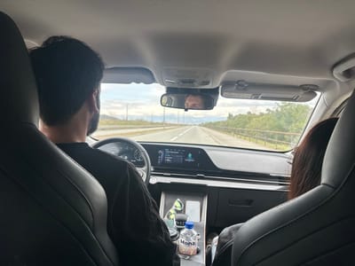 An interior view from the rear seat of a modern vehicle on a highway in Goričani, Montenegro. The driver, an adult male with dark hair and a beard, is visible from behind, wearing a dark shirt and operating the steering wheel. His eyes and forehead are reflected in the rearview mirror, indicating he is looking forward. To his right, an adult female front passenger with long dark hair is partially visible from behind. The car's dashboard features a large digital display across the instrument cluster and infotainment system. A "Mona" branded water bottle, containing colorful small objects, rests in a cup holder on the center console. The vehicle is traveling on a multi-lane highway, bordered by green foliage and a guardrail on the right, and distant hills under a partly cloudy sky on the left.