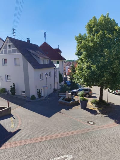 This image depicts a street scene in Hardthausen am Kocher, Germany. The perspective is from an elevated viewpoint, looking down at a residential street. In the foreground, a cobblestone pavement with red brick edging forms a circular pattern. To the left, a light grey residential building with a dark roof is visible. The building has multiple windows and a prominent pitched roof with dormer windows. Two small, sculpted evergreen trees are planted in front of this building.
Further into the scene, a street lamp with a sign reading "Brennweinstr. 3" is located at the corner of a street. Adjacent to the street lamp is a small water feature with stone pillars and a shallow basin. To the right of the water feature, a large, mature tree with dense green foliage dominates the right side of the image, casting shadows on the street. A dark grey SUV is parked near the water feature and the large tree, with a portion of a white car visible further down the street.
In the background, more residential buildings with red-tiled roofs are visible, along with a cellular tower on one of the rooftops. The sky is clear and blue, indicating a sunny day. The overall environment is a quiet residential area with paved streets and well-maintained buildings and landscaping.