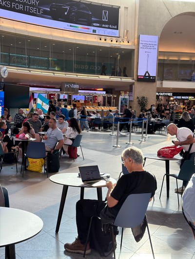 The image shows a busy airport waiting area. Several people are seated, some with laptops and bags, suggesting they are travelers waiting for flights. The scene is brightly lit, indicating it's daytime. The architecture of the airport is visible, with large windows and tiled floors.
Visible text includes advertisements and airport signage. One prominent advertisement promotes the iPhone, highlighting features like immediate color availability, up to 24 interest-free installments, and no interest or VAT. Another sign reads "AVATR-IL.CO.IL". There's also text related to "DUTY FREE" shopping.
The people in the image appear to be in a state of waiting. Their postures and the presence of luggage suggest travel. Some individuals are engaged with their devices, while others seem to be resting or conversing. The overall atmosphere appears to be one of casual waiting, common in airport environments. The flags visible, possibly representing different countries, add to the international context of an airport.