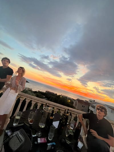 The image shows a sunset in Kruče - Kërruçi, Montenegro. Three people are visible on a balcony. One man wears sunglasses and a black shirt. A woman stands with a man in a black shirt; she wears a light-colored blouse and white pants. A table is covered with empty wine bottles, glasses, cigarettes, and a handbag. The sky shows a mix of grey clouds and orange from the setting sun above the sea. Buildings are visible beyond the balcony.