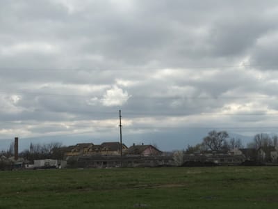 The image depicts a desolate rural landscape under a heavy overcast sky. In the foreground is a field of green grass. In the midground is a cluster of buildings, varying in condition. Some appear to be relatively new residences with yellow siding, while others are dilapidated structures with flat roofs. A tall brick chimney rises from the left side. An electrical pole is vertically positioned near the centre. Bare trees and sparse vegetation surround the buildings. In the background, a faint mountain range is discernible through the haze. Overlapping the top portion of the image is a series of electrical wires. The image does not contain any people. The location is likely on the outskirts of Sibiu, Romania, based on the architecture and geographical features.