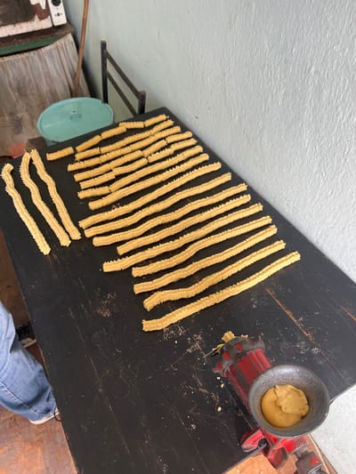 The image shows a tabletop in Hrishteni, Bulgaria, with numerous long, ridged, yellowish dough strips arranged in rows. A red, hand-cranked meat grinder-like device with a gray bowl filled with more dough is positioned on the right side of the table. A turquoise metal object, possibly a pan lid, sits on a shelf below the table, along with other miscellaneous items, including a green and white object. A section of a person's leg in blue jeans and a white sneaker is visible in the lower right corner. The wall in the background is light gray.