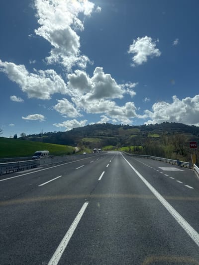 The image displays a multi-lane asphalt highway near Ancona, Italy, captured from a vehicle's forward perspective on a sunny day. The sky is bright blue with numerous scattered white cumulus clouds, and the sun is visible as a glare in the upper-left quadrant. The highway features white solid and dashed lane markings, along with faded yellow lines, and extends into the distance, curving gently to the right. Metal guardrails line both sides of the road. On the left side, a silver-colored van is parked or moving slowly on an adjacent service road or shoulder, set against a green grassy embankment. Further ahead on the main highway, several trucks are visible in the distance. On the right side, a red "SOS" sign is mounted on a pole near the shoulder. The background consists of rolling hills covered with dense green vegetation, including trees and grass. No people are discernible in the scene.