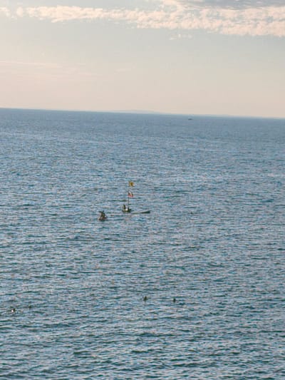 The image shows a wide expanse of calm, blue water under a hazy, pale sky with scattered white clouds. In the middle ground of the water, there are two small rafts or boats. One raft has a person sitting on it, and the other raft appears to have a person and a flag pole with flags. There are some other small, dark objects scattered in the water, which could be buoys or other debris. The overall scene depicts a tranquil moment on the water, possibly a recreational activity or a marker in the sea.