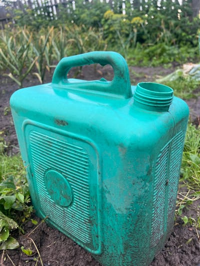 The image shows a dirty, green plastic jerrycan placed on the ground in an outdoor environment, likely a garden or yard. The jerrycan has a handle and an open spout. The ground is covered with soil, grass, and some dried leaves. In the background, there are green plants and a wooden fence. The overall impression is one of a functional item left outdoors in a slightly unkempt setting. The location is identified as Бучацінскі сельскі Савет, Belarus.
