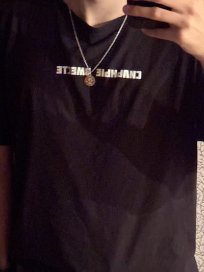 The image displays a chest-level, partial view of an individual indoors in Ciorescu, Moldova. The subject is wearing a black t-shirt with horizontally mirrored white text reading "CNUPHPIE 3WELLE" across the chest. A silver-colored chain necklace with a circular, gold-colored pendant featuring an intricate design hangs around the neck, resting on the t-shirt. Part of a hand is visible in the upper right quadrant, suggesting the individual is holding a camera or phone to capture the image. The background is dark and out of focus, with a narrow vertical strip of light-colored, patterned wallpaper visible along the far right edge of the frame.