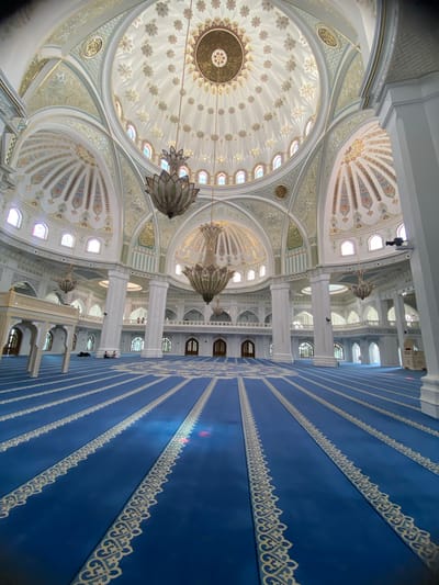 The image shows the interior of the Shali Grand Mosque, also known as the Akhmad Kadyrov Mosque, located in Shali, Russia. The mosque features a grand, ornate interior with a central dome and multiple smaller domes. The walls and ceilings are decorated with intricate patterns in gold and white, accented with pastel colors. Large chandeliers hang from the domes. The floor is covered with a vibrant blue carpet that is sectioned by white decorative borders, creating rows for prayer. In the foreground, the blue carpet stretches towards the back of the mosque, with rows of prayer markings visible. Several people are present in the mosque, appearing to be praying or resting on the carpet. The architecture suggests a large, significant religious building, with tall white columns supporting the arched structures and domes. The overall impression is one of grandeur and solemnity.