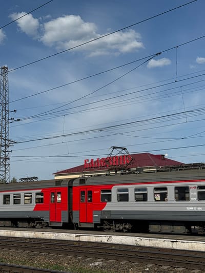 An outdoor shot of a red and grey passenger train with the Russian Railways (RZD) logo, stationary at a station in Balezino, Russia. The train's pantograph is visible, connected to overhead power lines against a cloudy blue sky. The number "3D4M 021606" is printed on the train car. Train tracks run along the platform edge.