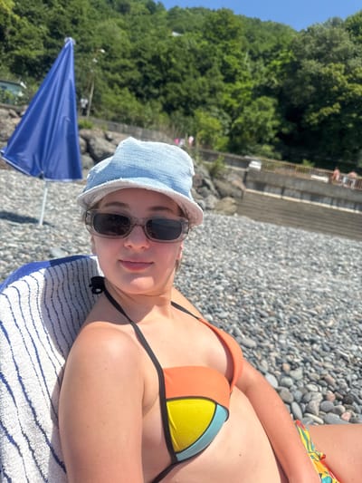 A young woman lies on a striped towel on a pebbled beach. She is wearing a light blue bucket hat and sunglasses, and her orange, yellow, and blue color-blocked bikini top is visible. Her right arm rests across her torso, and her left arm is bent at the elbow, partially obscuring her chest. The beach is covered in grey and white pebbles, and a blue umbrella is partially visible in the lower left corner of the frame. Lush green trees and a stone structure are visible in the background. The sun appears to be shining, casting shadows and highlighting the woman's skin. The scene evokes a relaxed, sunny day at the beach. The image is oriented in portrait mode.