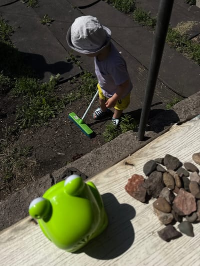 In an outdoor scene bathed in bright sunlight, a young child wearing a light-colored sun hat, a pale purple t-shirt, and bright yellow shorts is engaged in sweeping. The child is positioned on a dark, paved surface, likely a patio or pathway, with patches of green grass and soil interspersed. A long, dark metal pole runs across the upper left of the frame, casting a shadow. The child is holding a bright green broom with a long silver handle, actively sweeping the ground. Their feet are clad in black and white striped sandals.
To the right of the child, a collection of various-sized stones are arranged on a light-colored wooden surface. The wood appears weathered and has a distinct grain. In the foreground, a bright green frog-shaped toy with white circular eyes is visible. The frog toy is positioned on the wooden surface, and its shadow is cast onto the ground behind it.
The overall setting appears to be a residential backyard or garden area, given the presence of paving, grass, and a wooden structure. The strong shadows indicate it is likely midday or early afternoon on a sunny day. The child's focused posture while sweeping suggests they are occupied with a chore or play activity. No text is visible in the image. The location context is Зыково, Russia.