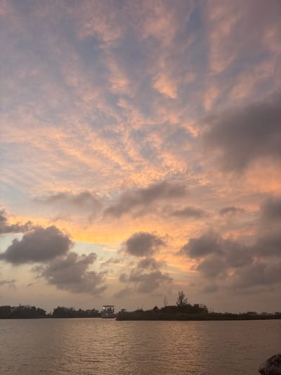 The image shows a body of water, likely a river or bay, with a treeline on the opposite bank. The sky is filled with clouds illuminated by the setting sun, creating a warm, orange glow. There is a boat or structure visible on the water near the treeline. The location is potentially near Tampico, Mexico, based on the visual information.