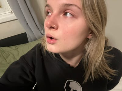A young woman lies on her side, partially facing the camera. She has light blonde hair, fair skin, and a silver nose ring. She wears a black sweatshirt with a white emblem on the upper chest. Behind her, a gray curtain and a folded green blanket are visible. The angle is slightly off-kilter, with the background appearing behind her head. The room is simple, with white walls showing minimal detail.