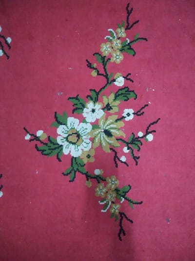 The image displays a close-up, overhead view of a patterned floor covering located in Chimay, Belgium. The dominant background color is solid red, exhibiting a textured appearance and scattered small white specks, consistent with dust or minor surface wear on a textile or linoleum material. A stylized floral design is integrated into this red background. The central motif consists of dark, thin, branching stems from which dark green, angular leaves emanate. Flowers and buds are depicted along these branches. The main floral elements include larger blossoms featuring white petals with yellow-green centers, outlined in black. Smaller clusters of flowers or buds are also present, predominantly yellow-green with white details and black outlines. Very small white dots represent additional buds along finer branches. Portions of identical floral patterns are visible at the periphery of the frame, indicating a repeating design. No individuals, actions, objects beyond the floor covering, or specific environmental context are discernible within the frame.