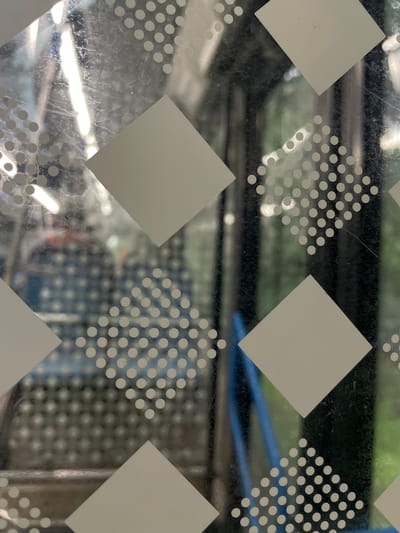 The image displays a close-up view of a glass surface with a diamond-patterned design. The pattern consists of large, solid white diamonds interspersed with clusters of small white dots arranged in diamond shapes. The glass appears to be part of a window or screen, as a blurred background is visible through it. The background shows hints of an indoor or outdoor environment with various objects, including what seem to be shelves, possibly containing items, and some greenery, suggesting a setting like a store, greenhouse, or an area with plants. There are also horizontal lines and a blue element in the background, which could be part of shelving or furniture. The lighting suggests it is daytime, with some bright reflections on the glass, indicating artificial or natural light sources. The overall scene is abstract due to the focus on the patterned glass and the blurred background. There is no discernible text in the image.