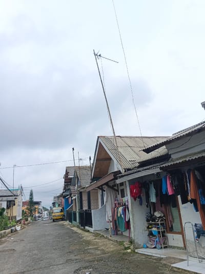 The image presents a residential street in Kabiufa, Papua New Guinea, under an overcast sky. The asphalt road, showing signs of wear, leads uphill from the foreground. Buildings, primarily single-story structures with corrugated metal roofs and painted facades, line both sides of the street.
On the immediate right, a house features an outdoor area where various items of clothing, including shirts, pants, and dresses of multiple colors, are suspended on drying racks and lines. Adjacent to this, a multi-tiered shoe rack holds several pairs of footwear, and a blue child's scooter is positioned on the ground. A tall, slender pole extends vertically from this structure, supporting a television antenna with visible lead-in wires.
Utility lines are strung across the street between poles. Further down the street on the left, a yellow box truck or mini-bus is parked. A small, conical evergreen tree is visible in the mid-ground on the left. No individuals are present in the scene.
