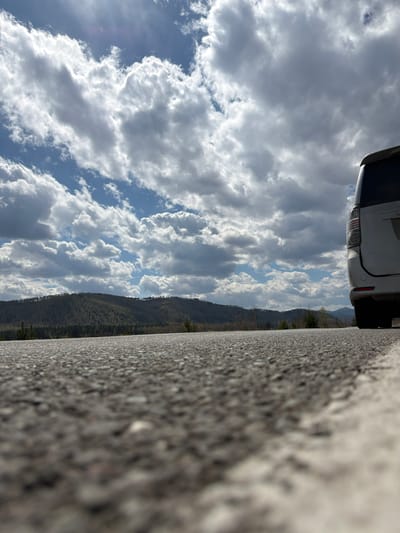 The image depicts a low-angle perspective of a scene that includes a road, a distant mountain range, a cloudy sky, and part of a white car. The foreground is dominated by a coarse, asphalt road surface. A mountain range with dense green vegetation stretches horizontally across the midground. The sky is filled with fluffy white clouds with patches of blue visible in between. The rear of a white car is visible. Based on the current image data, no conclusions can be made about Сизая, Russia.