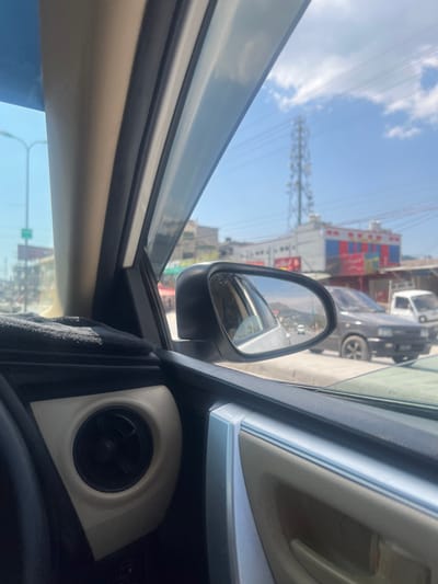 This image captures a street scene in Abbottabad, Pakistan, from the interior perspective of a vehicle's front passenger seat. The vehicle's cabin displays beige and black trim, including a circular air vent in the lower left, a black fabric object on the dashboard, and a light-colored A-pillar.
Outside the vehicle, the environment is brightly lit under a blue sky with scattered white clouds. A prominent cellular tower is visible in the mid-background, with utility lines crossing the sky. Along the street, commercial buildings are present, one featuring a multi-colored facade (red, blue, white) with what appears to be Urdu signage. On the road, a dark-colored sedan is positioned to the right of the viewer's vehicle, with a white pickup truck behind it. The side mirror reflects the road extending into the distance, showing additional vehicles and distant mountains. A green road sign is partially visible on the left side of the street.