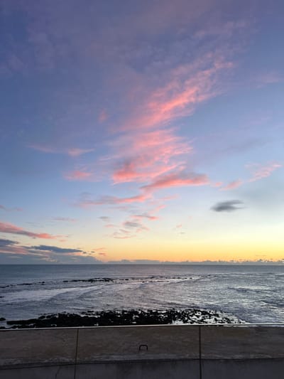 A sideways view of a sunset over the ocean. Pink-tinged clouds streak across a blue and yellow sky. The ocean stretches along the horizon, fading from gold to gray. A stone wall lines the right edge of the frame, with a rocky shoreline below. The overall scene captures the serene beauty of a coastal sunset.