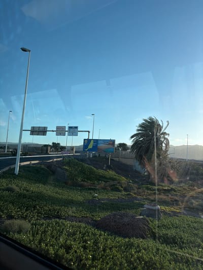 The image shows a landscape in Puerto del Rosario, Spain, observed through a window. A palm tree stands next to the road. Street signs indicate directions to Morro Jable, Gran Tarajal, Puerto del Rosario, Antigua, La Oliva, and Corralejo. A billboard advertises Fuerteventura. The environment features a mix of greenery and mountainous terrain in the background. The sky is clear and blue.